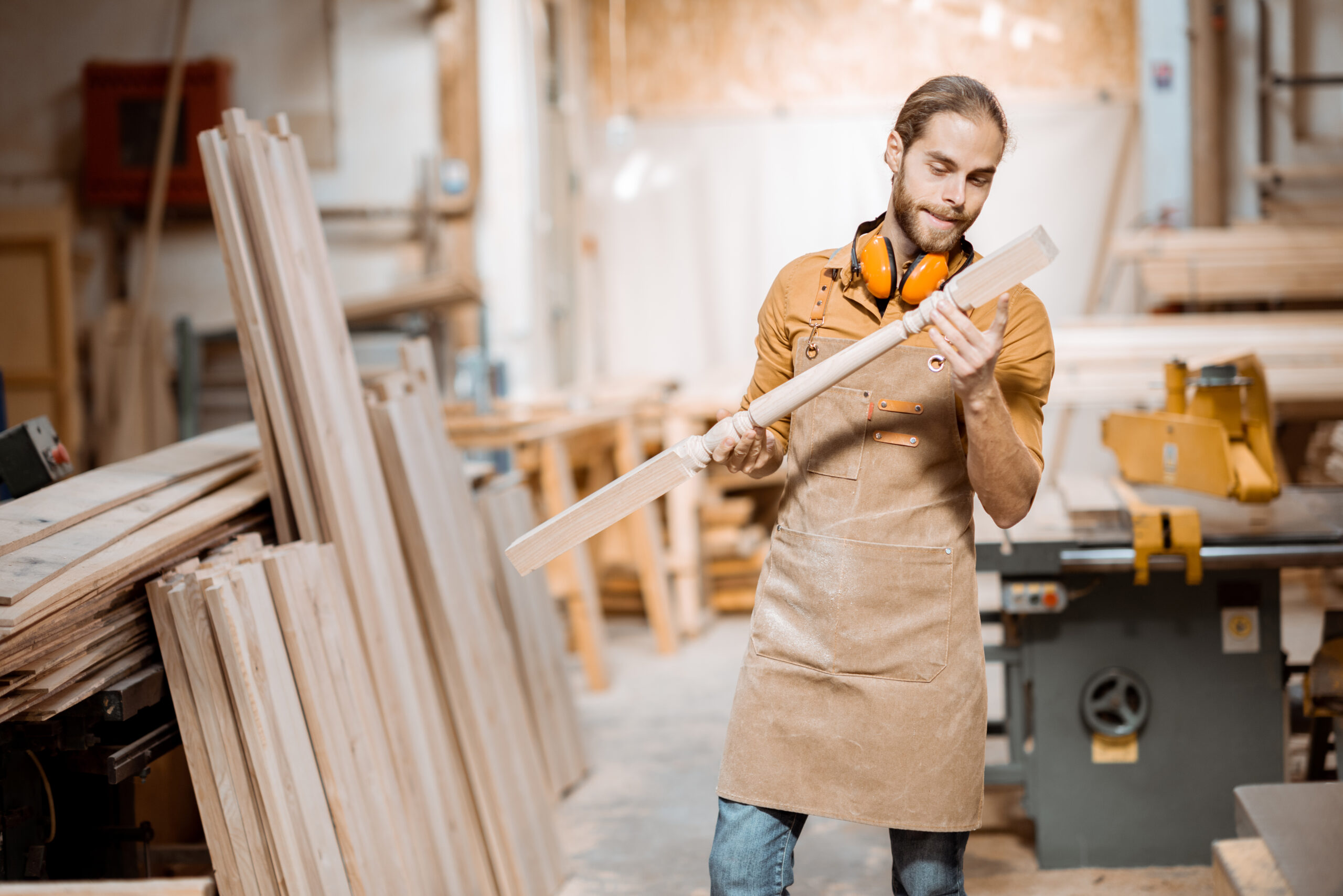 Handsome carpenter in uniform working with wood, checking the quality of the wooden baluster at the joinery
