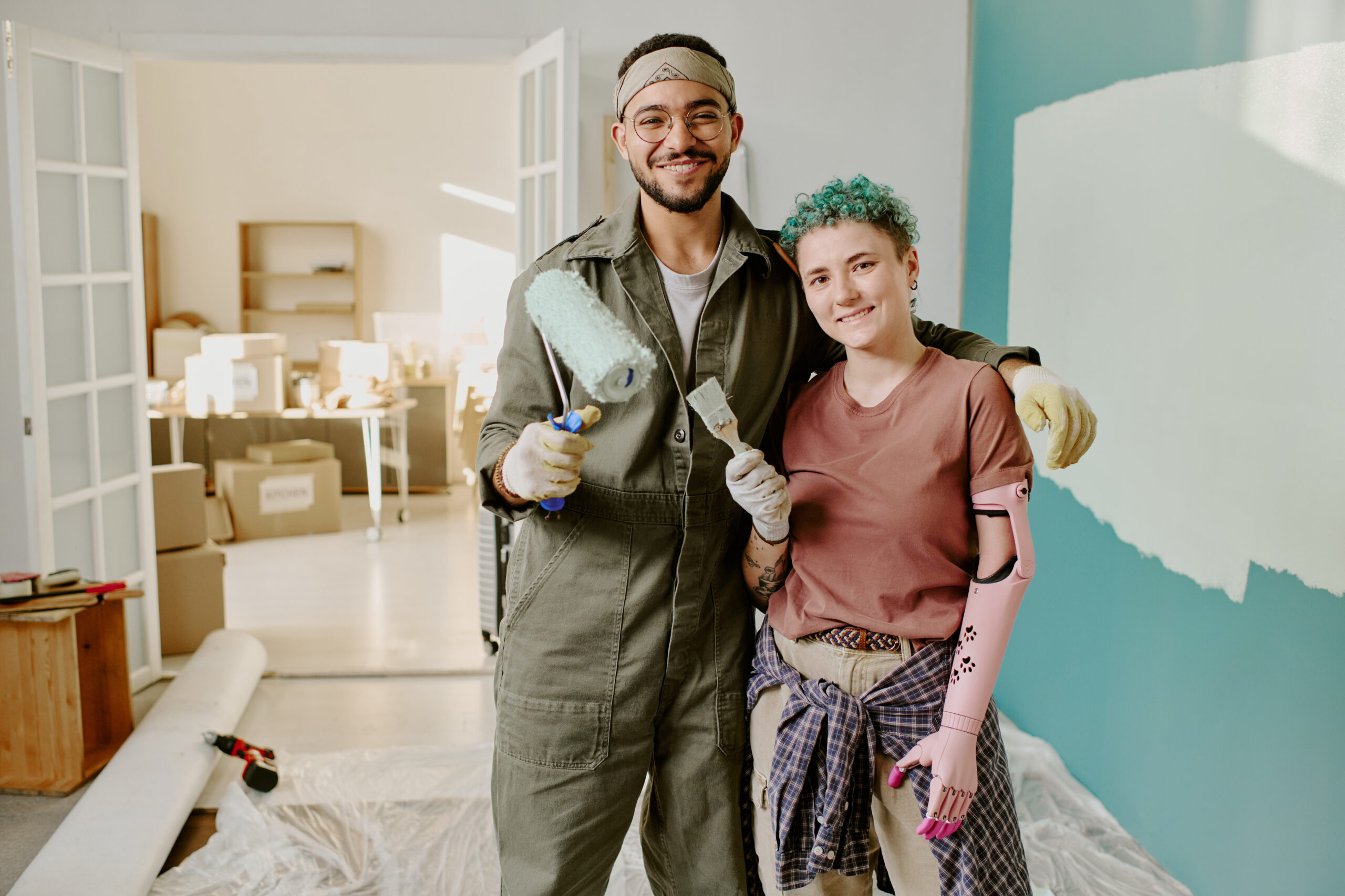 Young adult man and young adult Caucasian woman with prosthetic arm smiling while painting wall together during home renovation, standing close in bright room with moving boxes