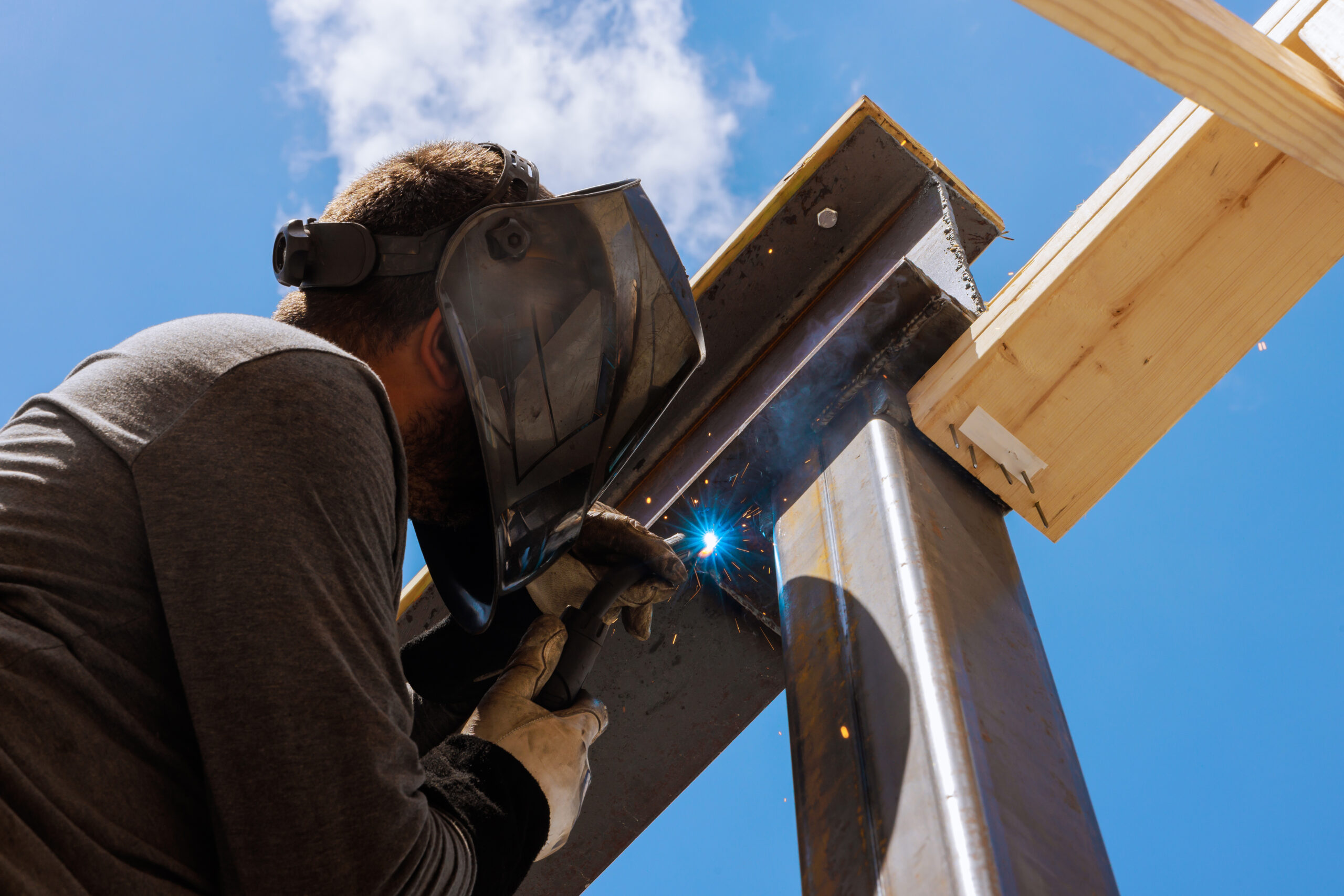 Contractor welds steel beams together at construction site, focusing intently on task while sparks fly.