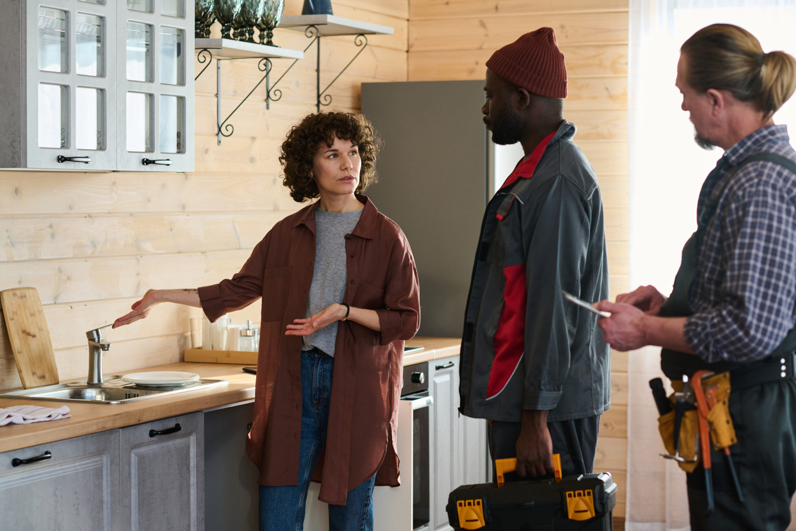 Young housewife opening metallic faucet and explaining problem to intercultural male technicians in workwear standing in front of her